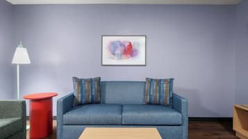 A living room with a blue sofa, two patterned pillows, a red side table, a floor lamp, and a framed abstract painting on the wall.
