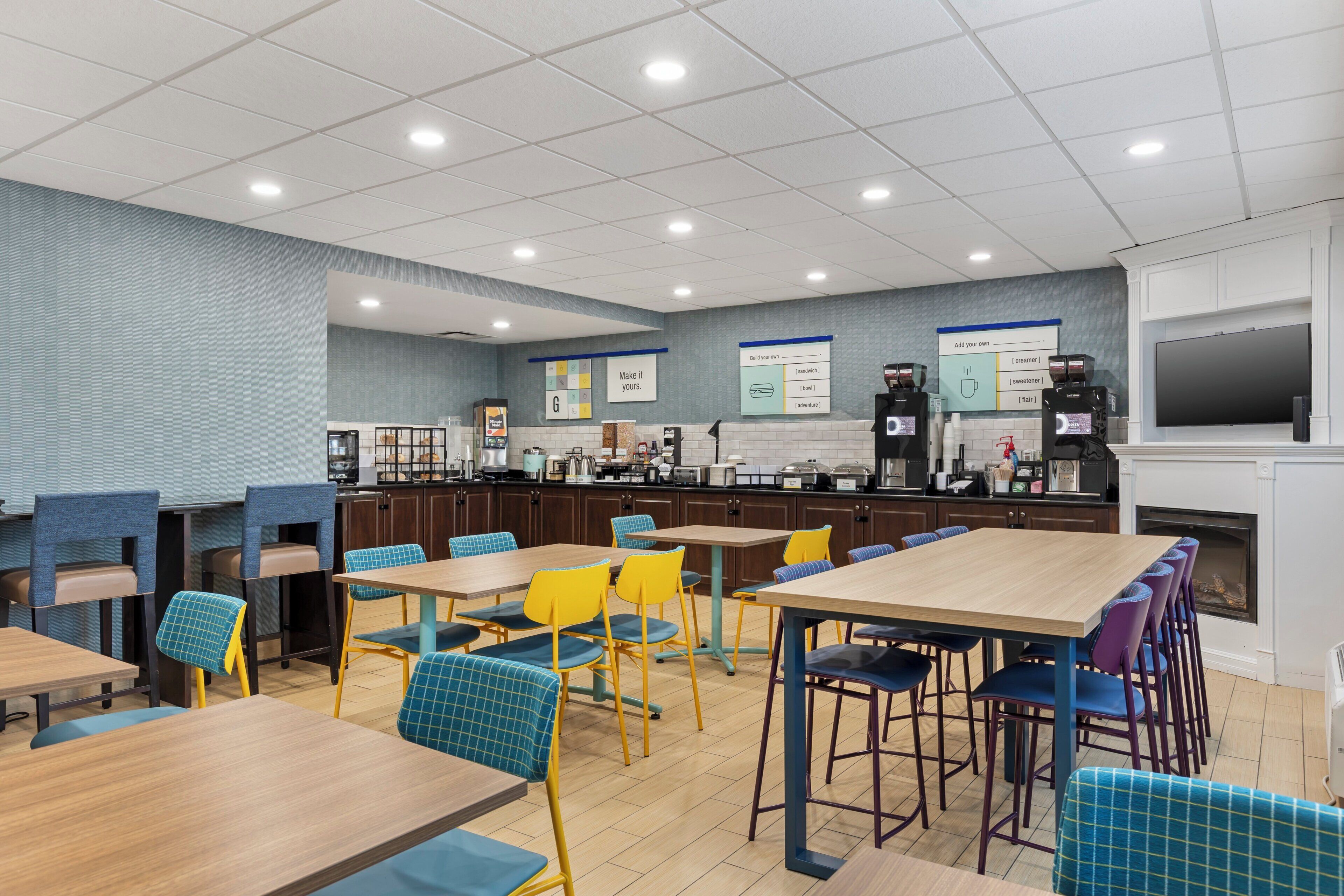 A modern cafeteria with multiple tables and chairs, a coffee station, and a television screen.