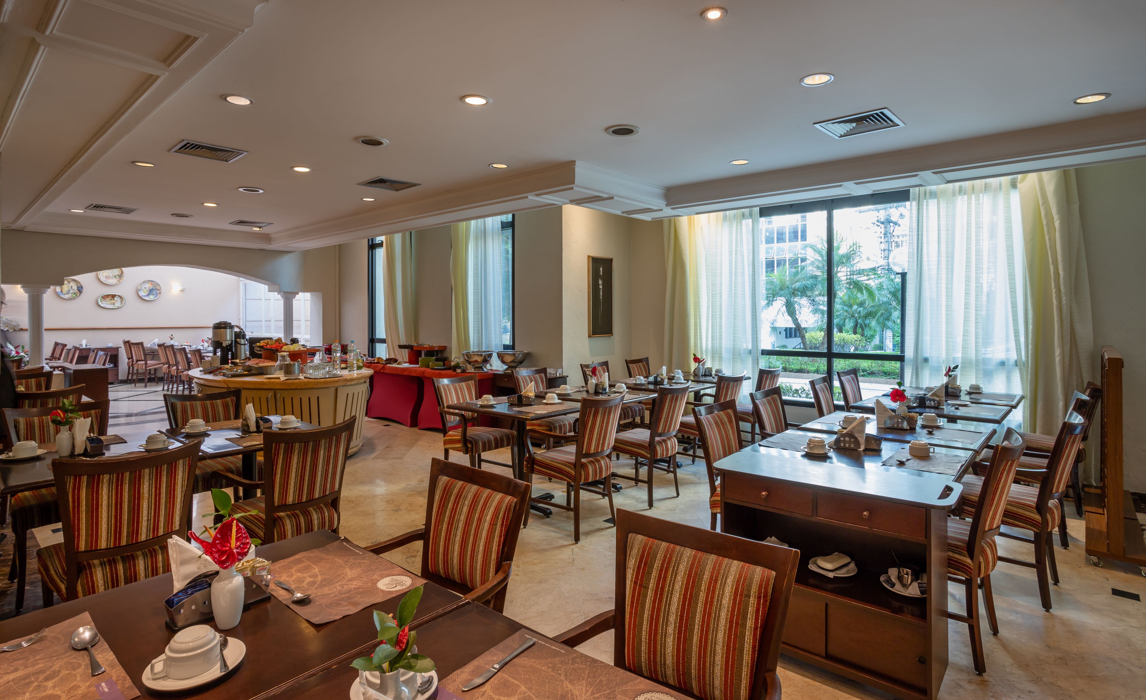A spacious dining area with multiple tables and chairs, a buffet setup, and large windows offering a view of the outdoors.