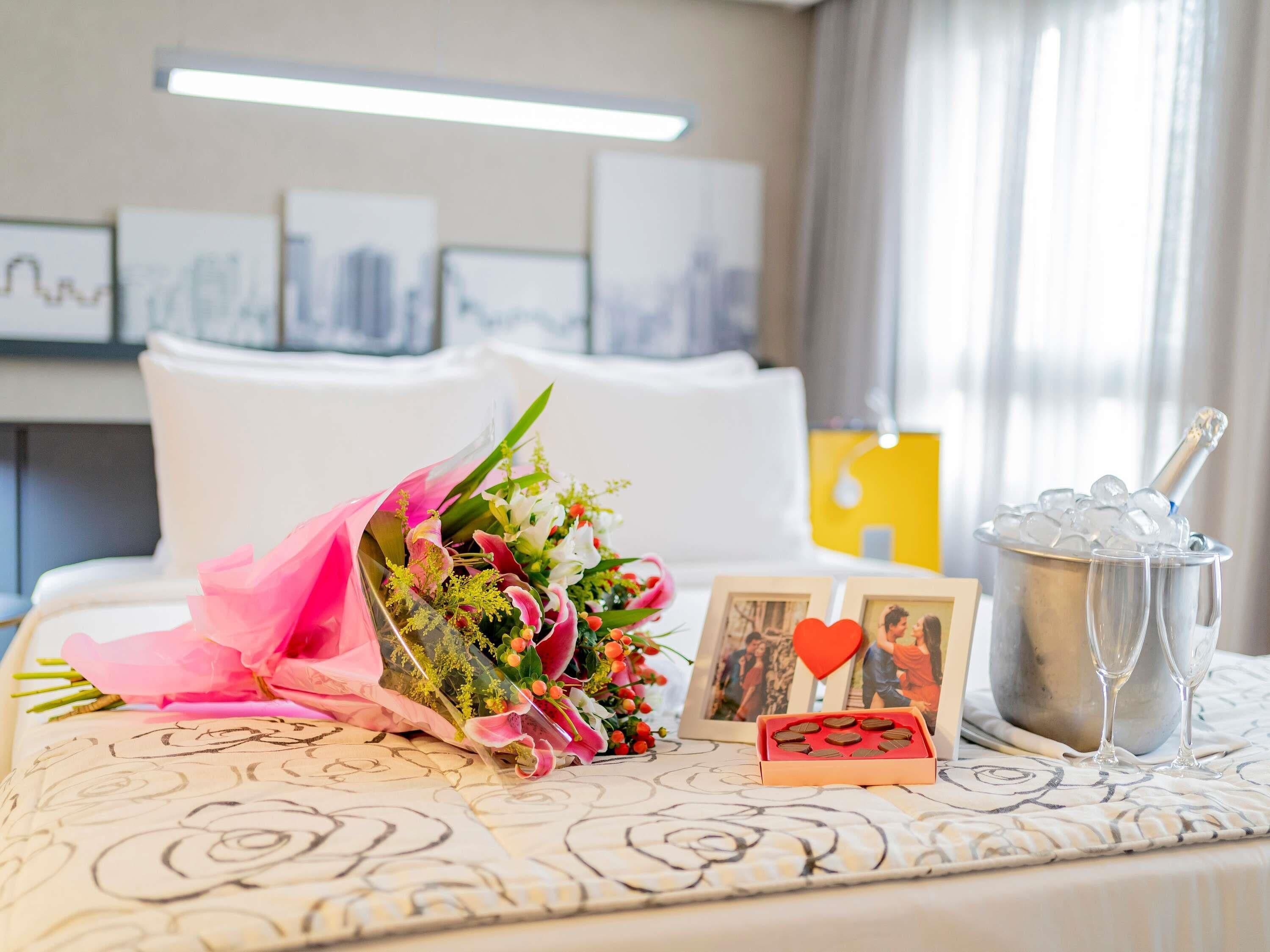 A neatly arranged hotel room with a bed, a bouquet of flowers, a photo frame, a small box of chocolates, and a champagne bucket.