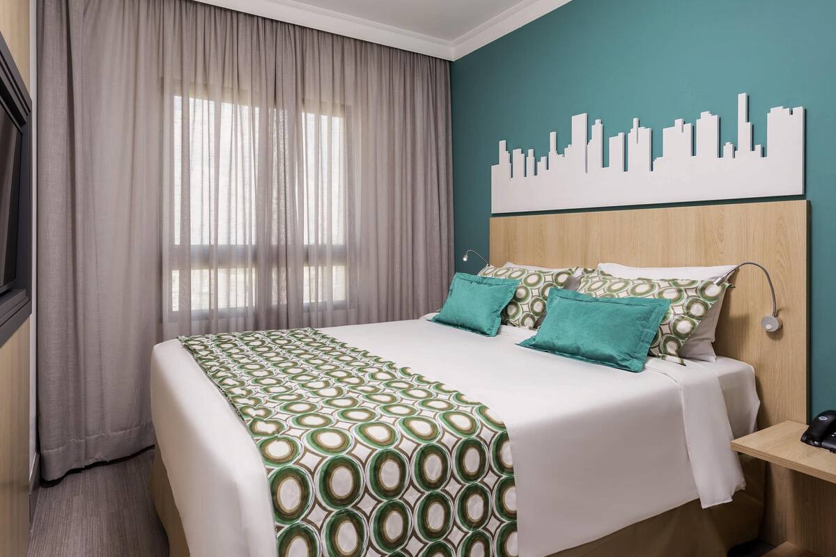 Een hotelkamer met een bed, gordijnen, stadsgezicht kunst aan de muur en een nachtkastje.