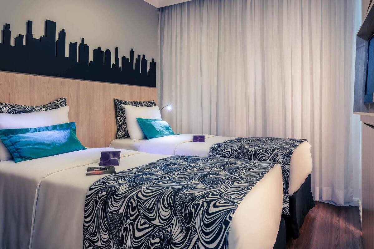 Een hotelkamer met twee bedden, een muurschildering van een stadszicht en een groot raam met gordijnen.
