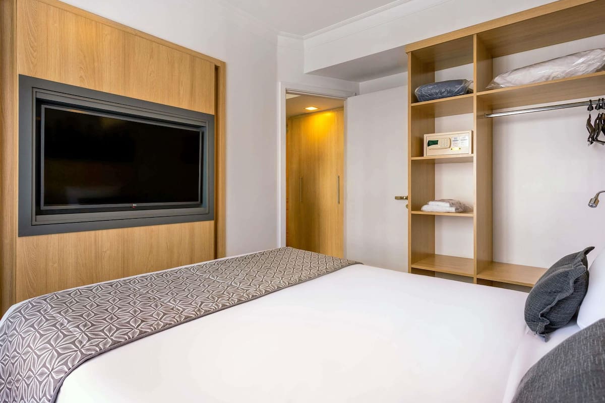 Una habitación de hotel con cama, televisión montada en la pared, armario y estante con objetos.
