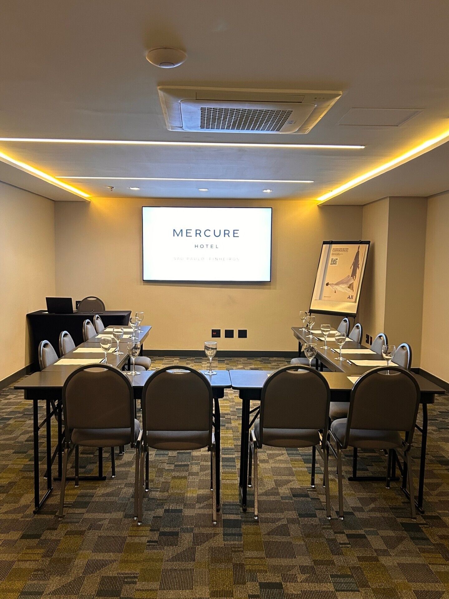 A conference room with a large screen displaying 'Mercure Hotel' and 'São Paulo, Brazil'. The room has a carpeted floor, a long table with chairs, and a projector screen.