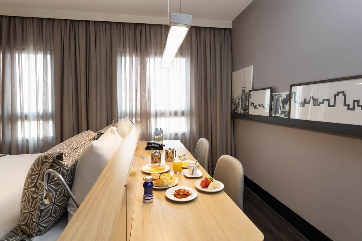 Quarto de hotel com uma longa mesa de jantar, um sofá, uma pintura com paisagem urbana e uma cama com almofadas estampadas.