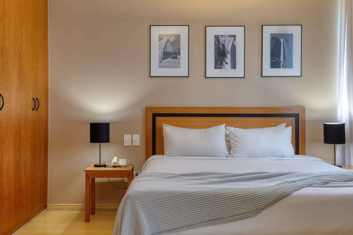 Habitación de hotel con una cama grande, un armario de madera, una mesita de noche con una lámpara y tres cuadros en la pared.