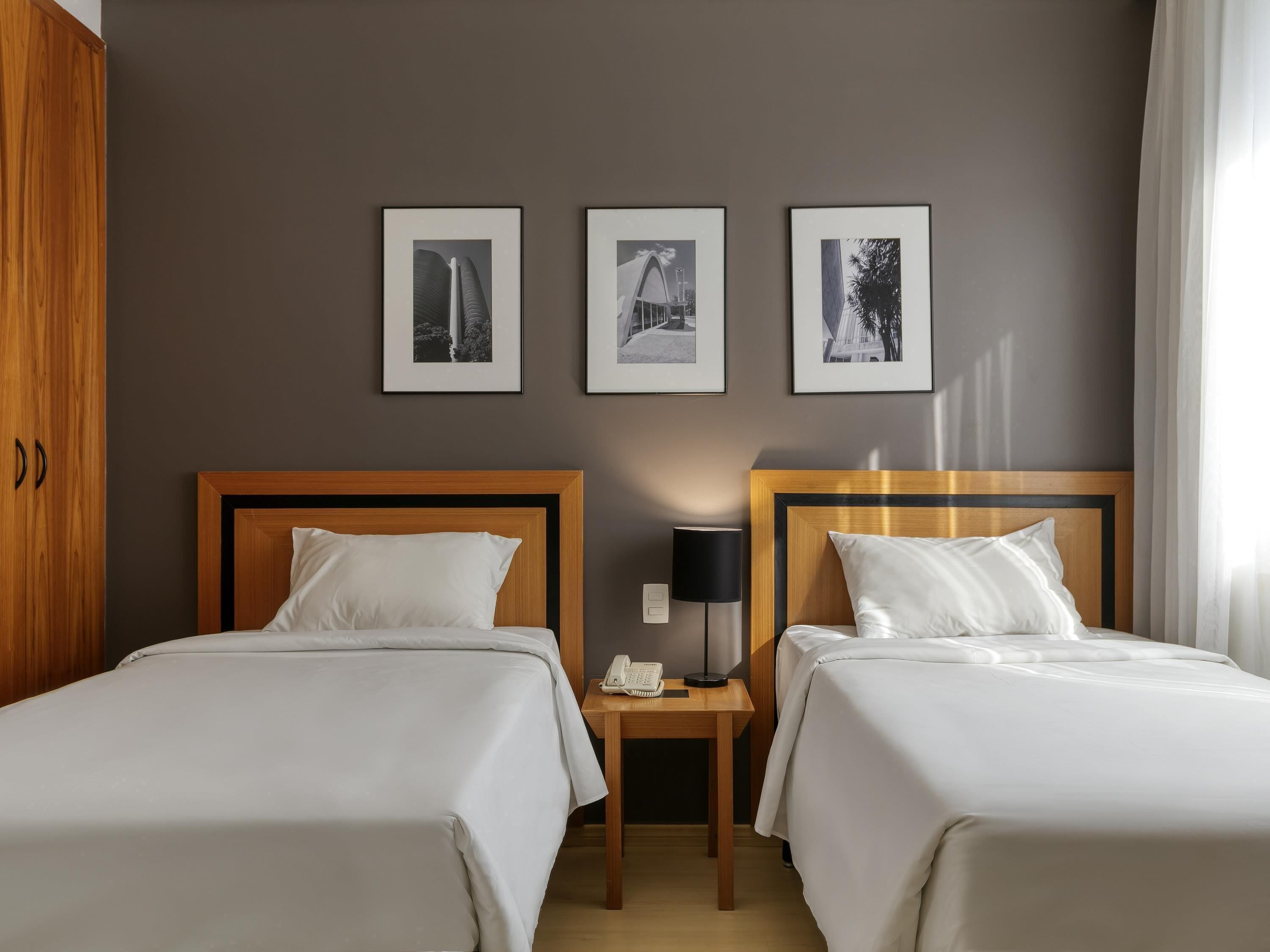 Una habitació d'hotel amb dos llits, un armari de fusta, una tauleta de nit amb una làmpada i tres quadres al mur.