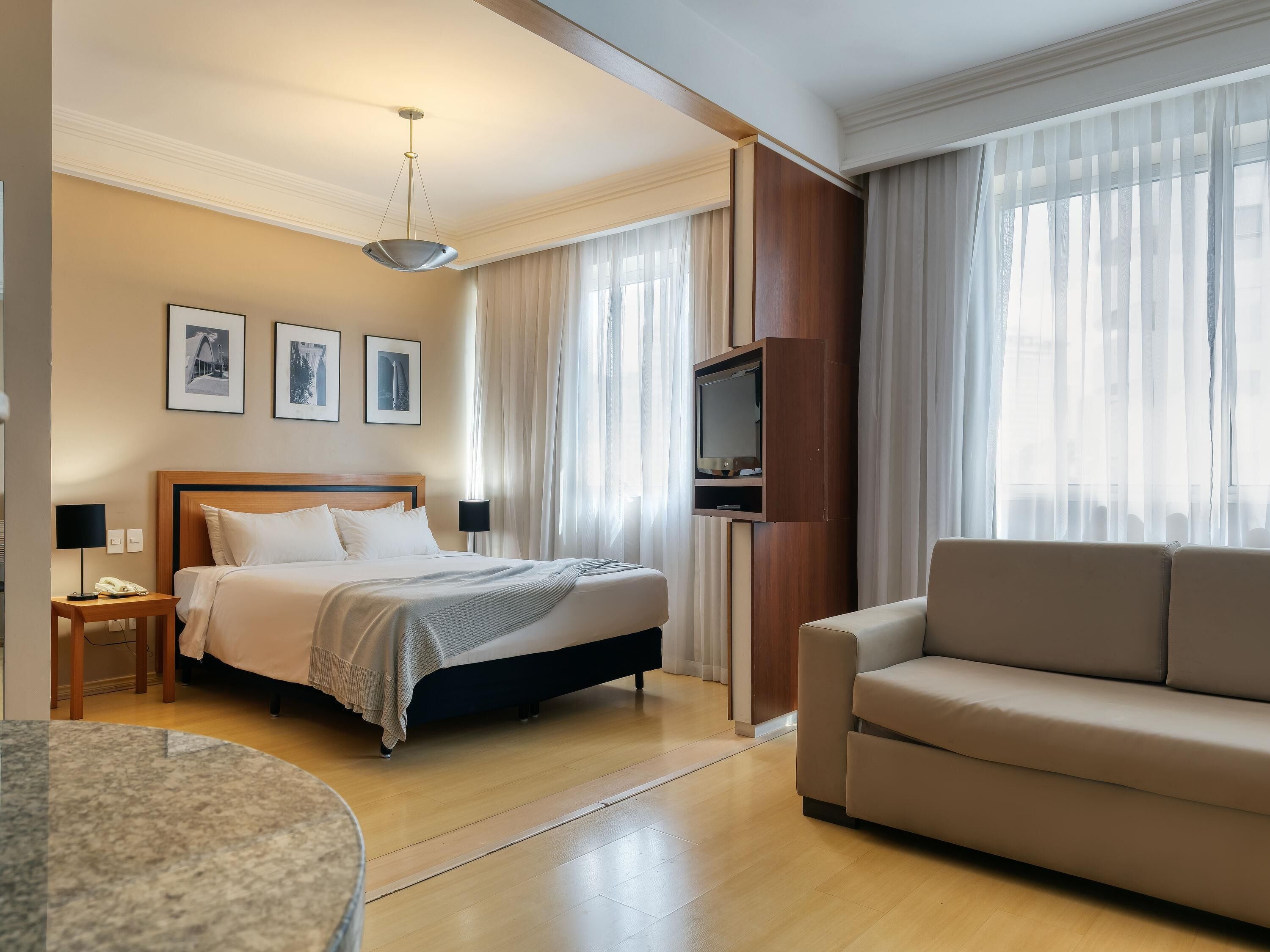 Una habitació d'hotel amb un llit, un sofà, un televisor fixat a un panell de fusta i quadres amb marc a la paret.
