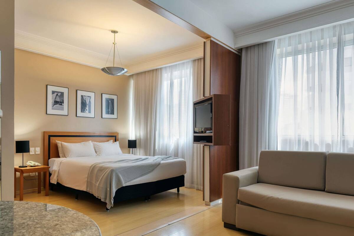 Una habitación de hotel con cama, televisión instalada en un panel de madera, sofá, una mesita y una lámpara.