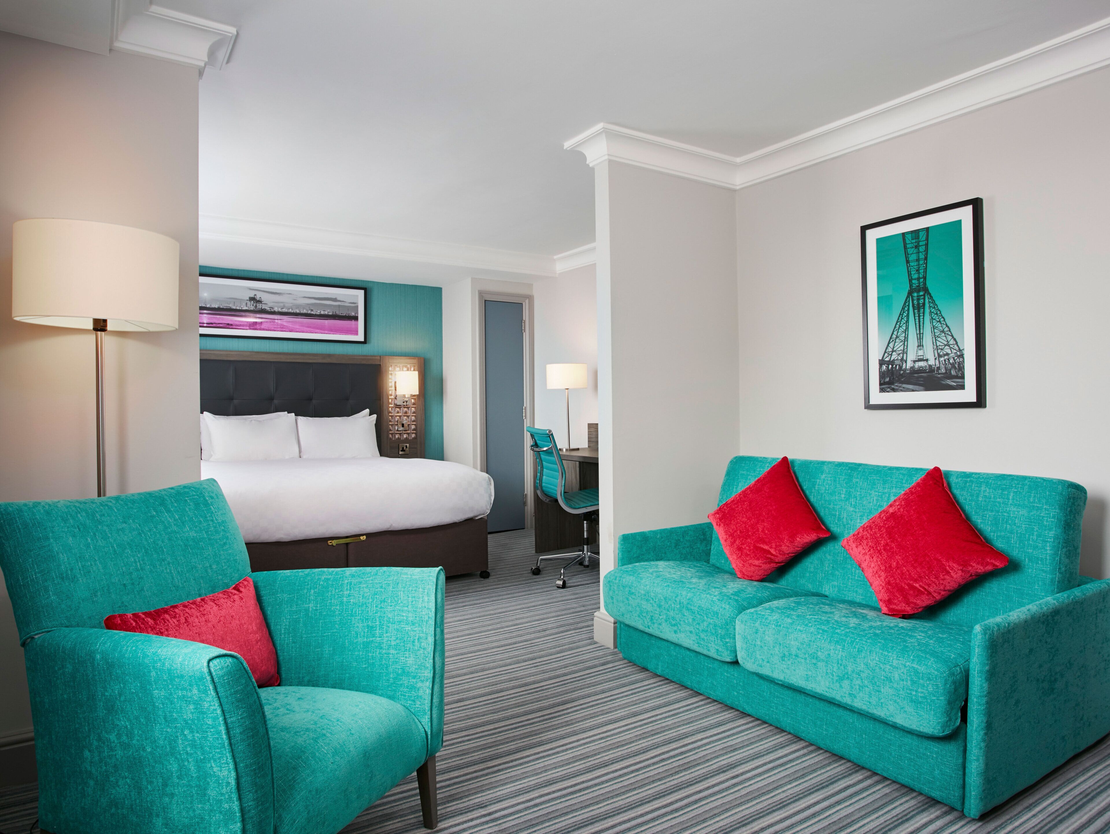 Una habitación de hotel con una cama, dos sillones turquesa, un sofá turquesa con cojines rojos y un cuadro enmarcado de una torre, además de una lámpara.