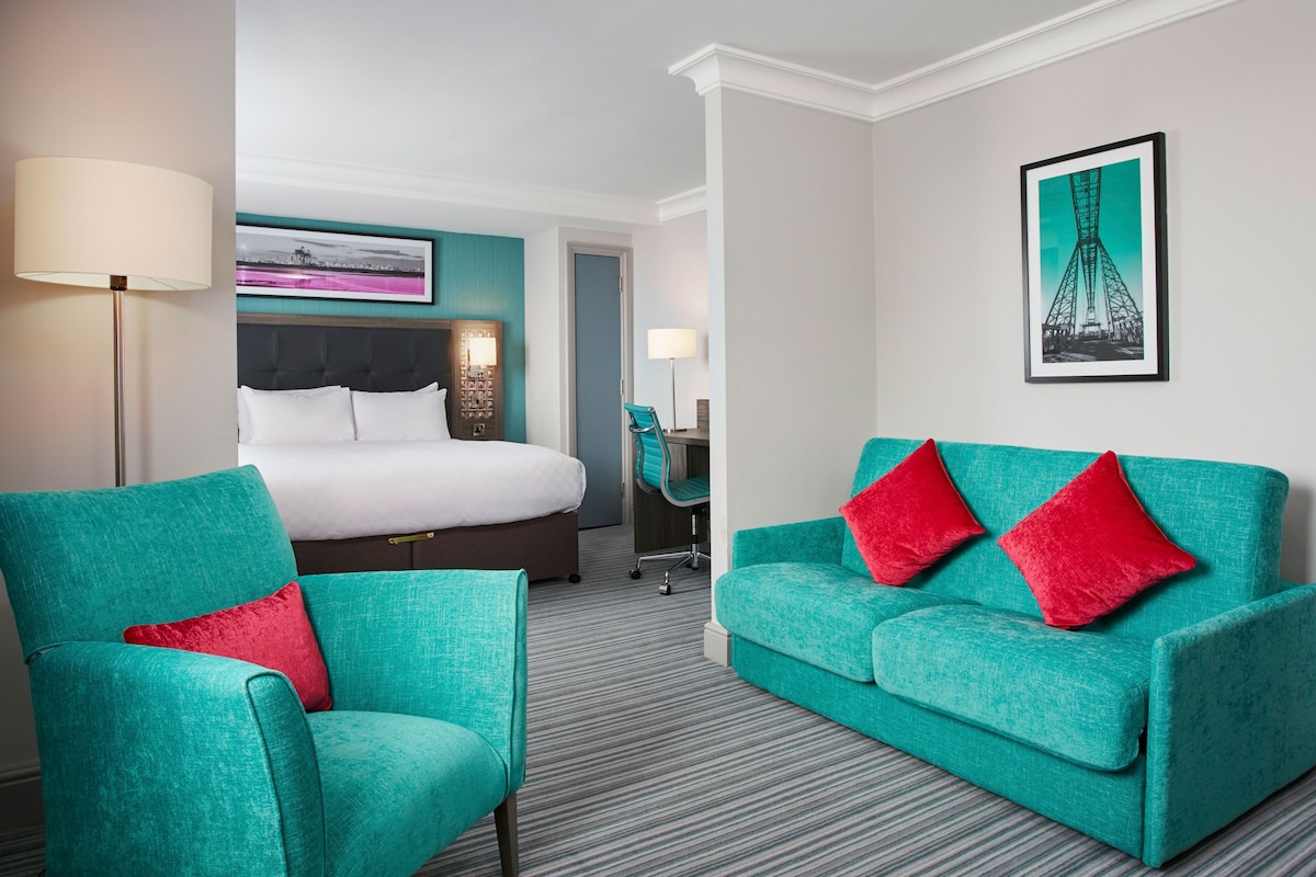 A hotel room with a bed, two teal armchairs, a teal sofa with red cushions, a framed picture of a tower, and a lamp.