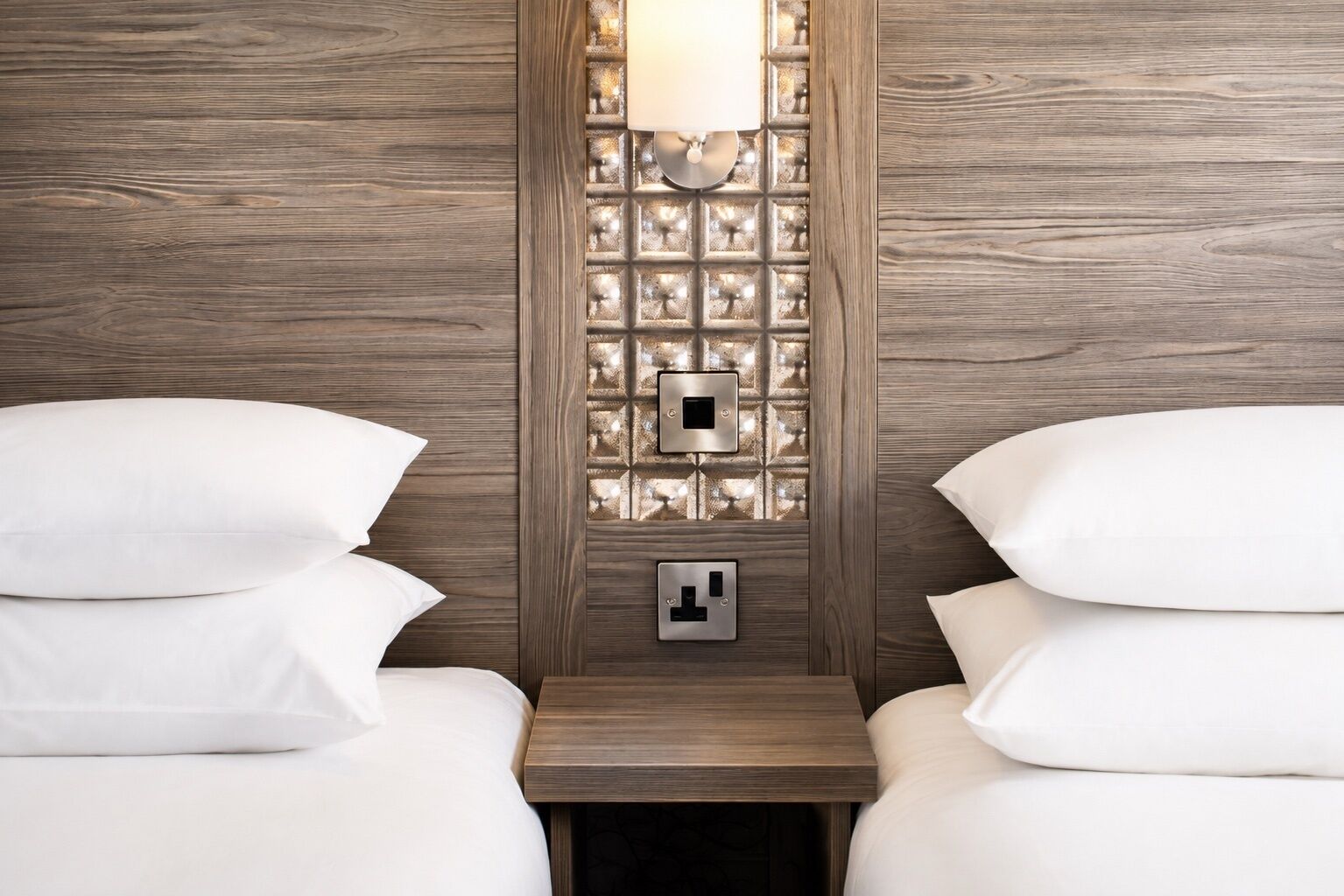 A hotel room with two beds, a nightstand, and a wall-mounted light fixture.
