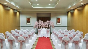 A hotel conference room with rows of white chairs with pink sashes, a red carpet aisle, and floral arrangements at the front.
