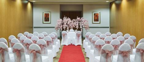 A hotel conference room with rows of white chairs with pink sashes, a red carpet aisle, and floral arrangements at the front.