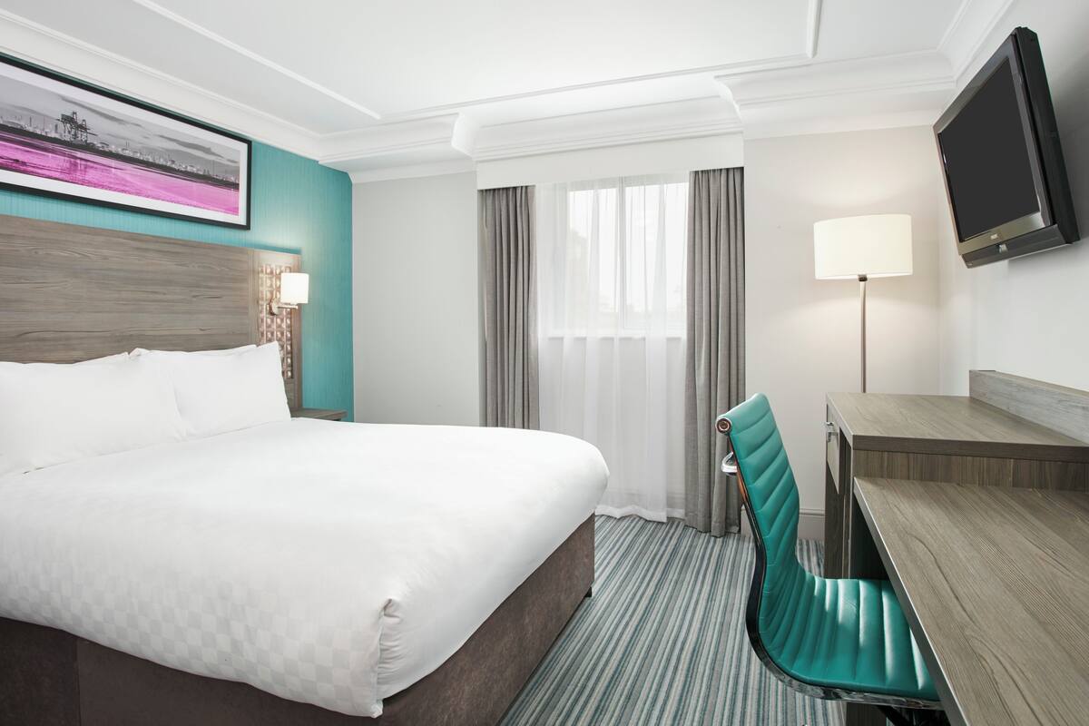 Una habitación de hotel con cama, escritorio, silla, televisor y ventana con cortinas.
