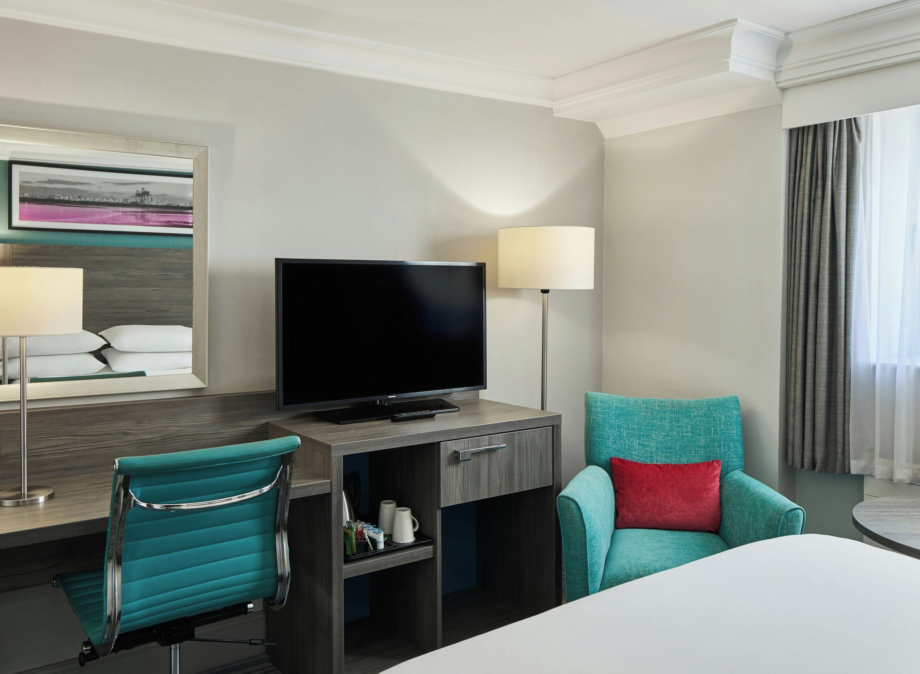A hotel room with a bed, a desk with a television, a chair, a lamp, and a window with curtains.