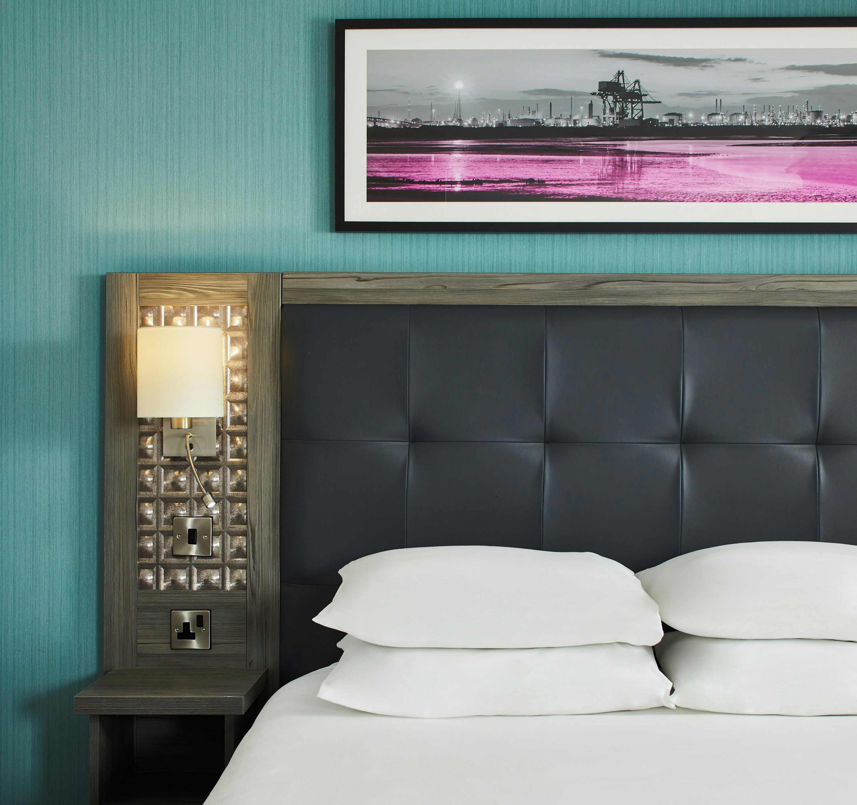 A hotel room with a dark headboard, a bedside table with a lamp, and a framed picture on the wall.