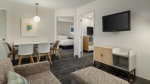 A hotel room with a sofa, a dining table with chairs, a TV mounted on the wall, and a bed in the background. - TownePlace Suites by Marriott Detroit Dearborn (Dearborn)