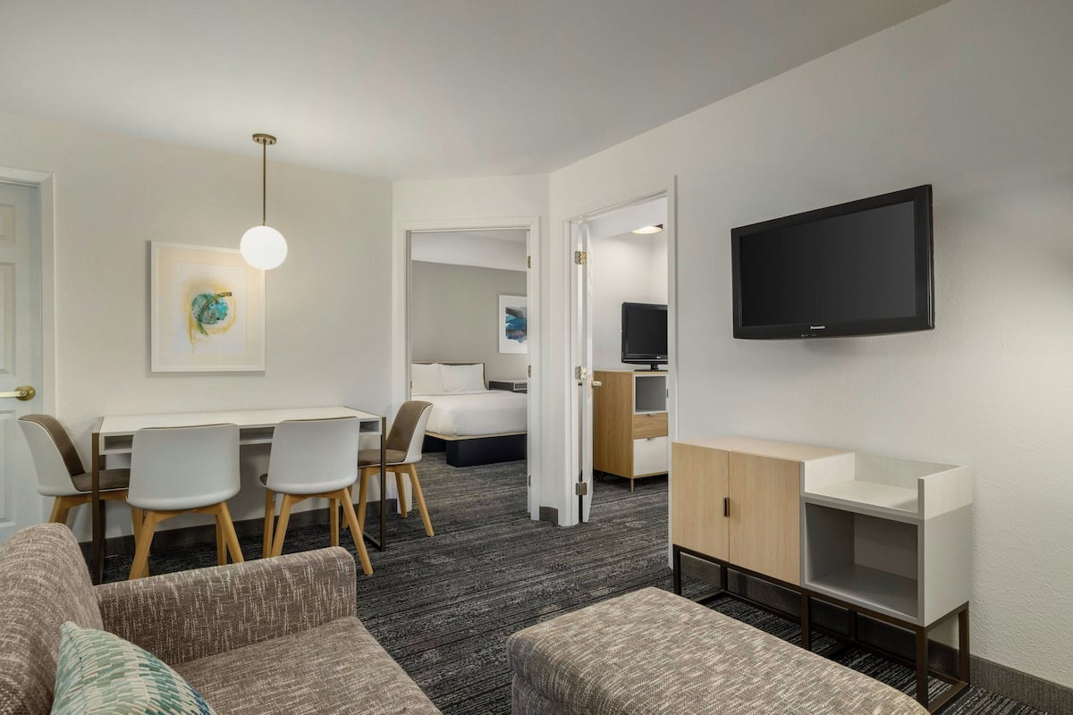A hotel room with a sofa, a dining table with chairs, a TV mounted on the wall, and a bed in the background.