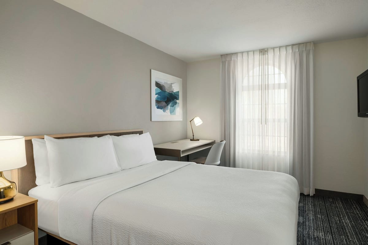 A hotel room with a large bed, a desk with a lamp, a chair, and a TV on the wall.