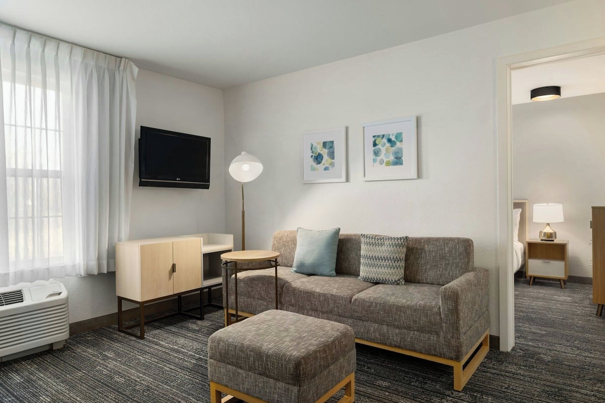 A hotel room with a sofa, ottoman, and a small table. There is a TV mounted on the wall, a lamp, and a window with curtains.