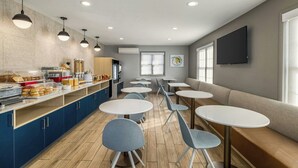 A modern cafe with round tables and blue chairs, a buffet counter with food items, and a TV mounted on the wall. - TownePlace Suites by Marriott Detroit Dearborn (Dearborn)