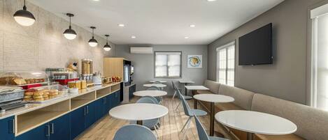 A modern cafe with round tables and blue chairs, a buffet counter with food items, and a TV mounted on the wall.