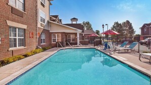 A rectangular pool with clear blue water, surrounded by a brick building with windows and a covered seating area with umbrellas. - TownePlace Suites by Marriott Detroit Dearborn (Dearborn)