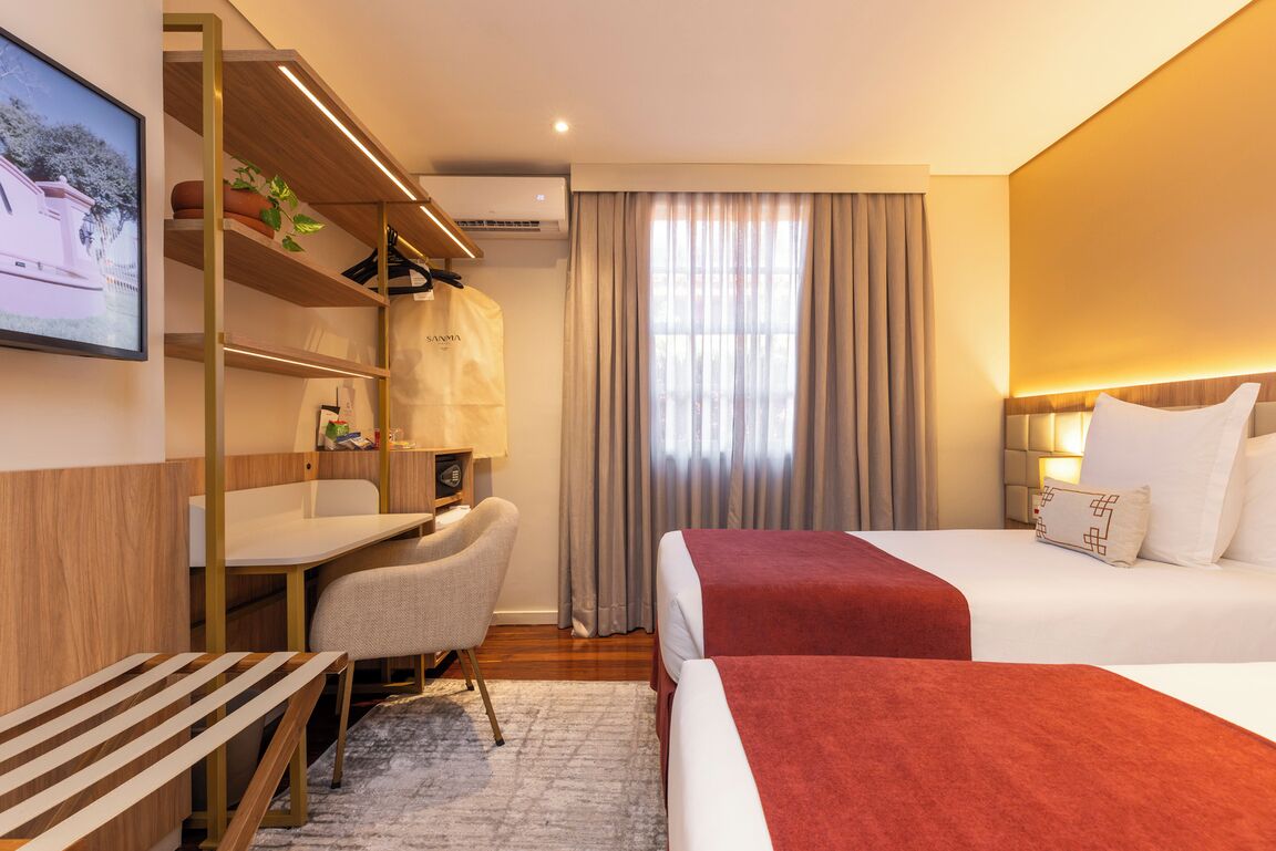 A hotel room with a bed, a desk, a chair, a shelf with a plant, and a window with curtains.