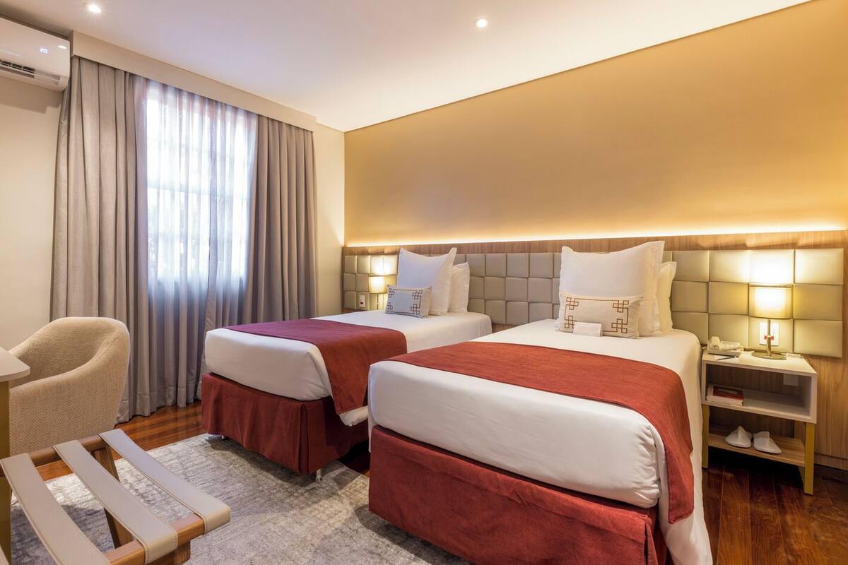 Quarto de hotel com duas camas, uma cadeira, uma mesinha de cabeceira e uma janela com cortinas.