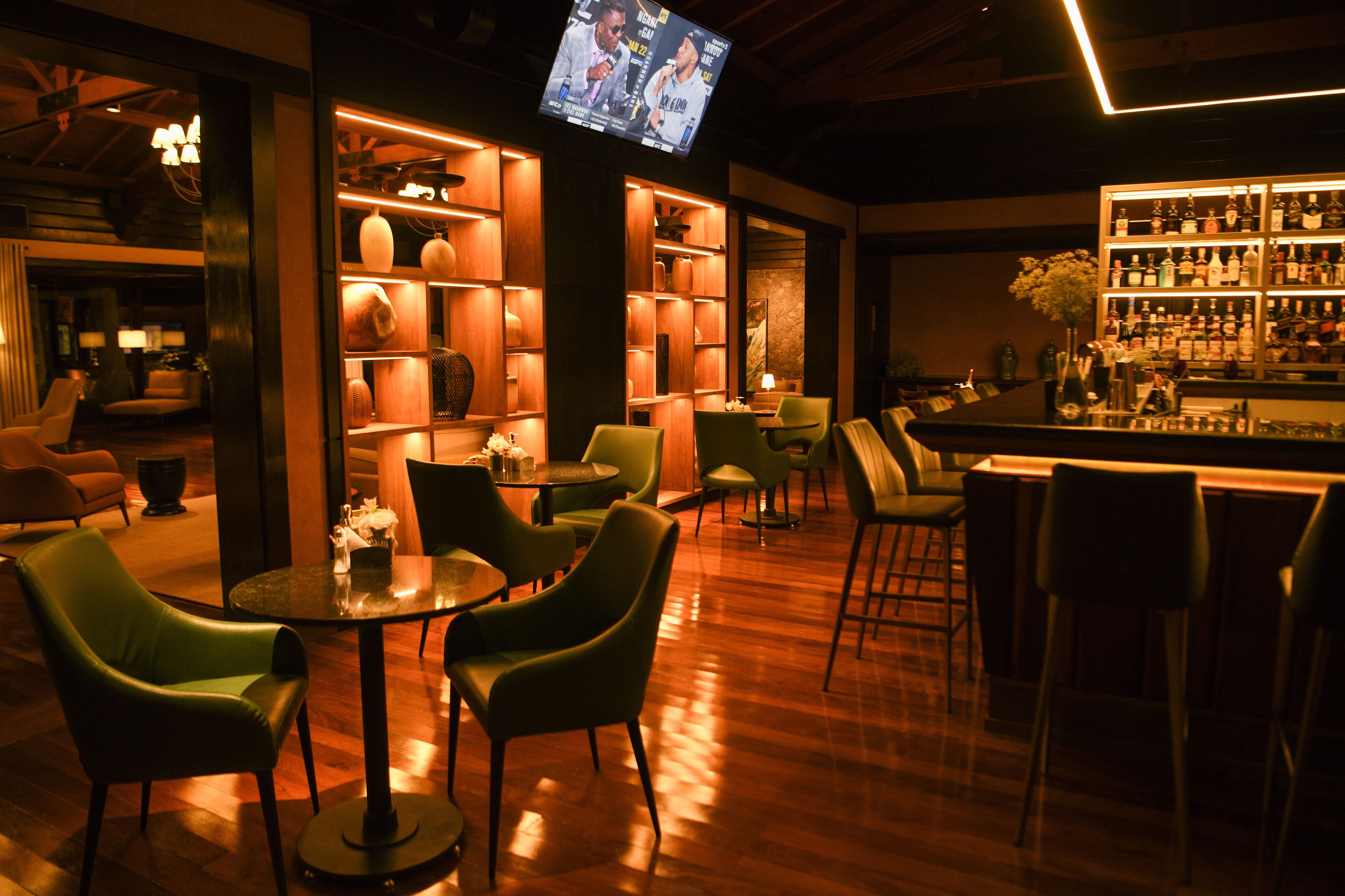 Una zona de bar con estanterías de madera, sillas verdes y una pantalla de televisión.