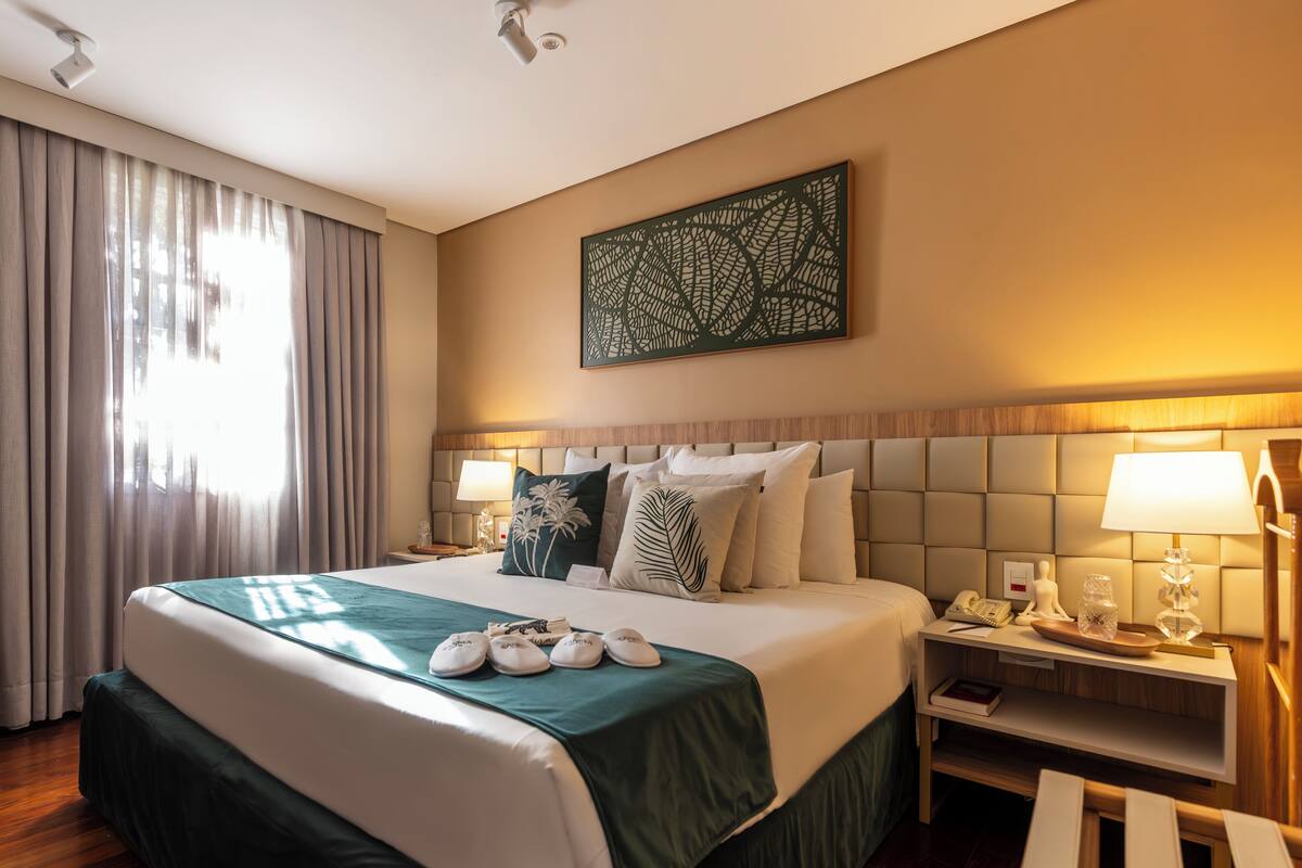 Quarto de hotel com uma cama grande, mesa de cabeceira, luminária e um objeto decorativo na parede.
