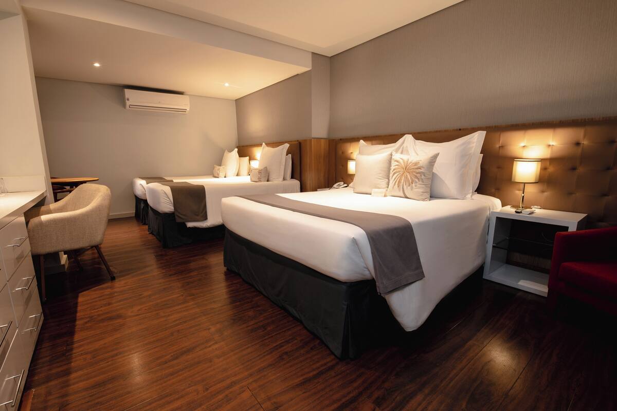 Quarto de hotel com duas camas, uma escrivaninha, uma cadeira e uma mesa de cabeceira com abajur.