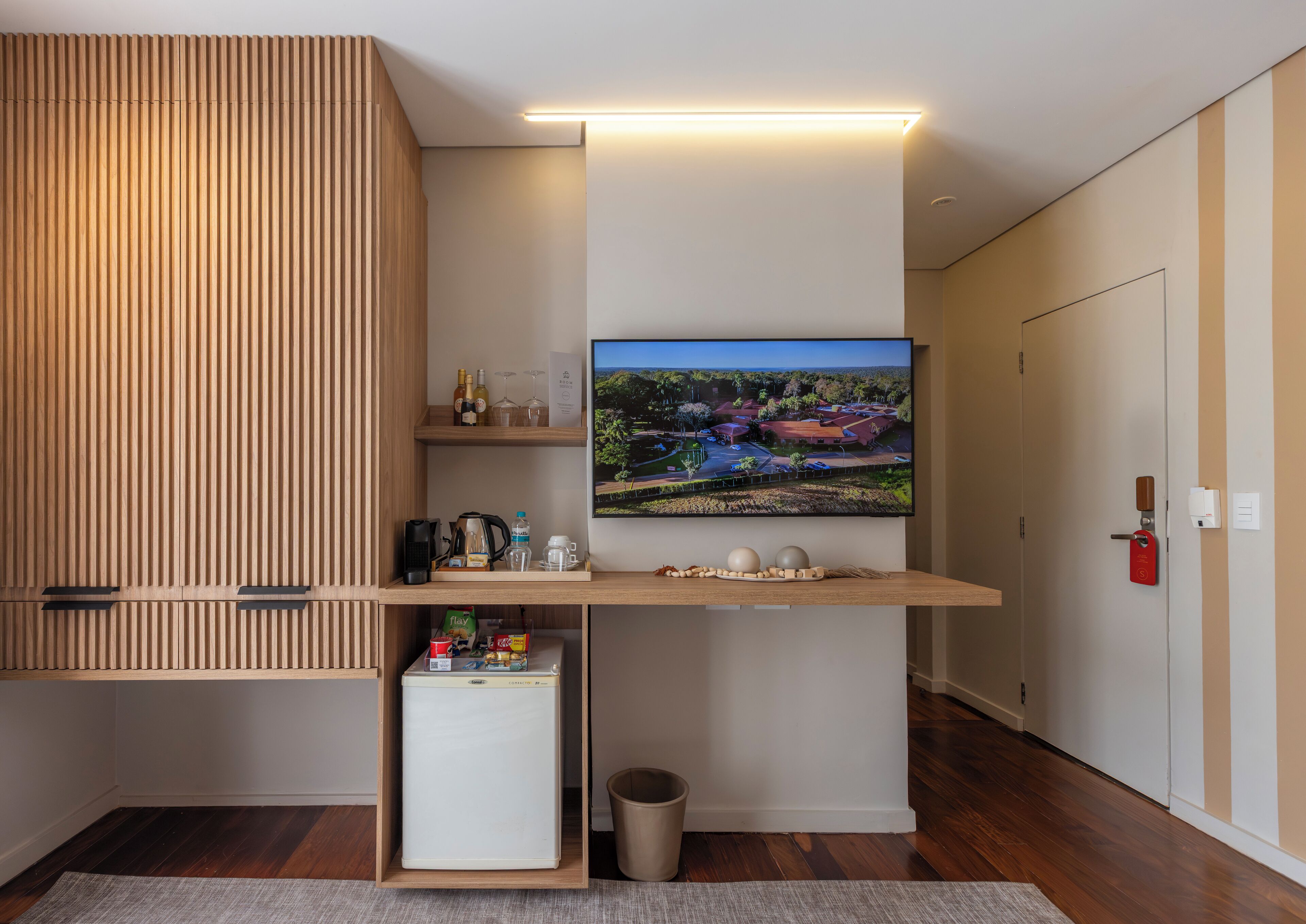 Una sala moderna con un televisor de pantalla plana montado en un estante de madera, un refrigerador pequeño y un basurero.