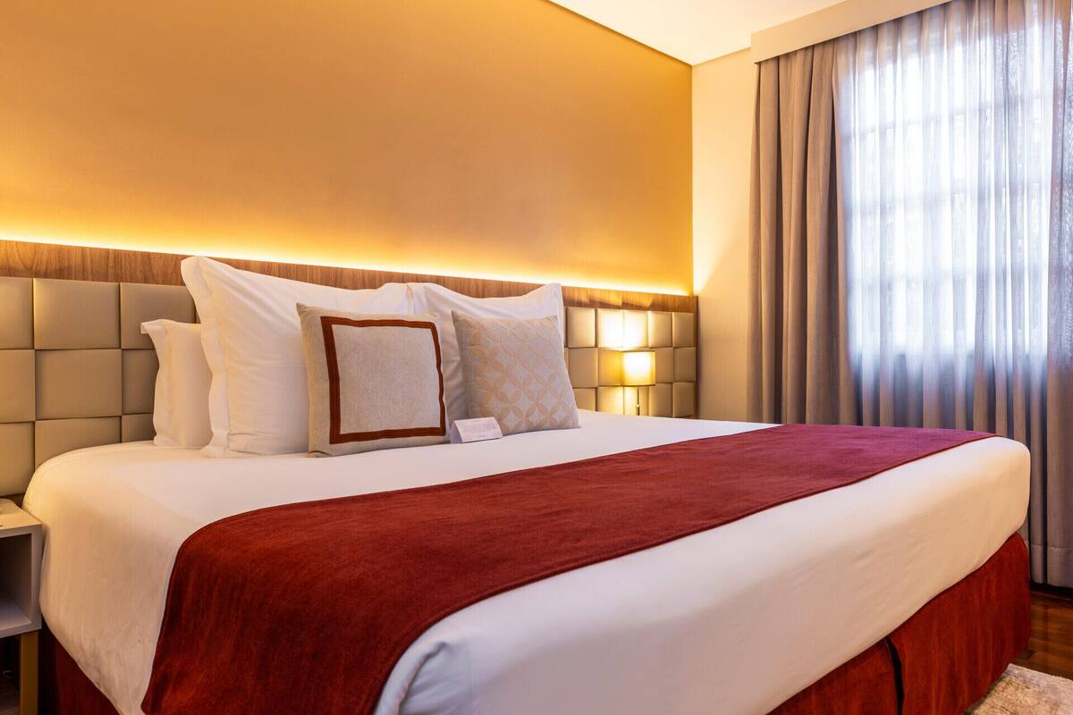 Quarto de hotel com uma cama grande, cabeceira com iluminação integrada, mesa de cabeceira e janela com cortinas.