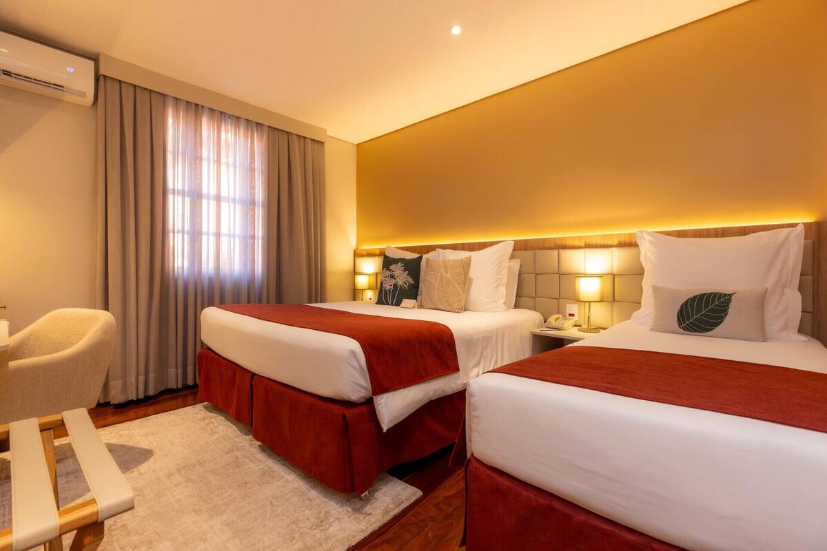 Quarto de hotel com duas camas, uma cadeira, uma escrivaninha e um ar condicionado.