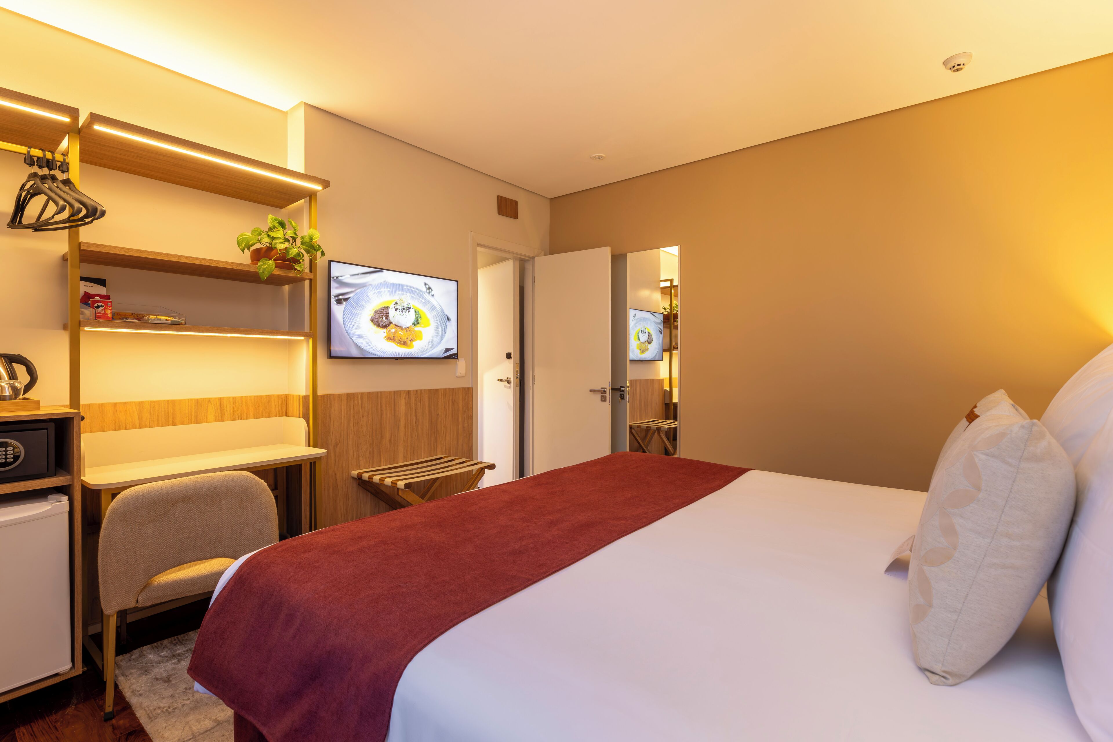 A hotel room with a bed, a desk, a chair, a TV, and a small plant.