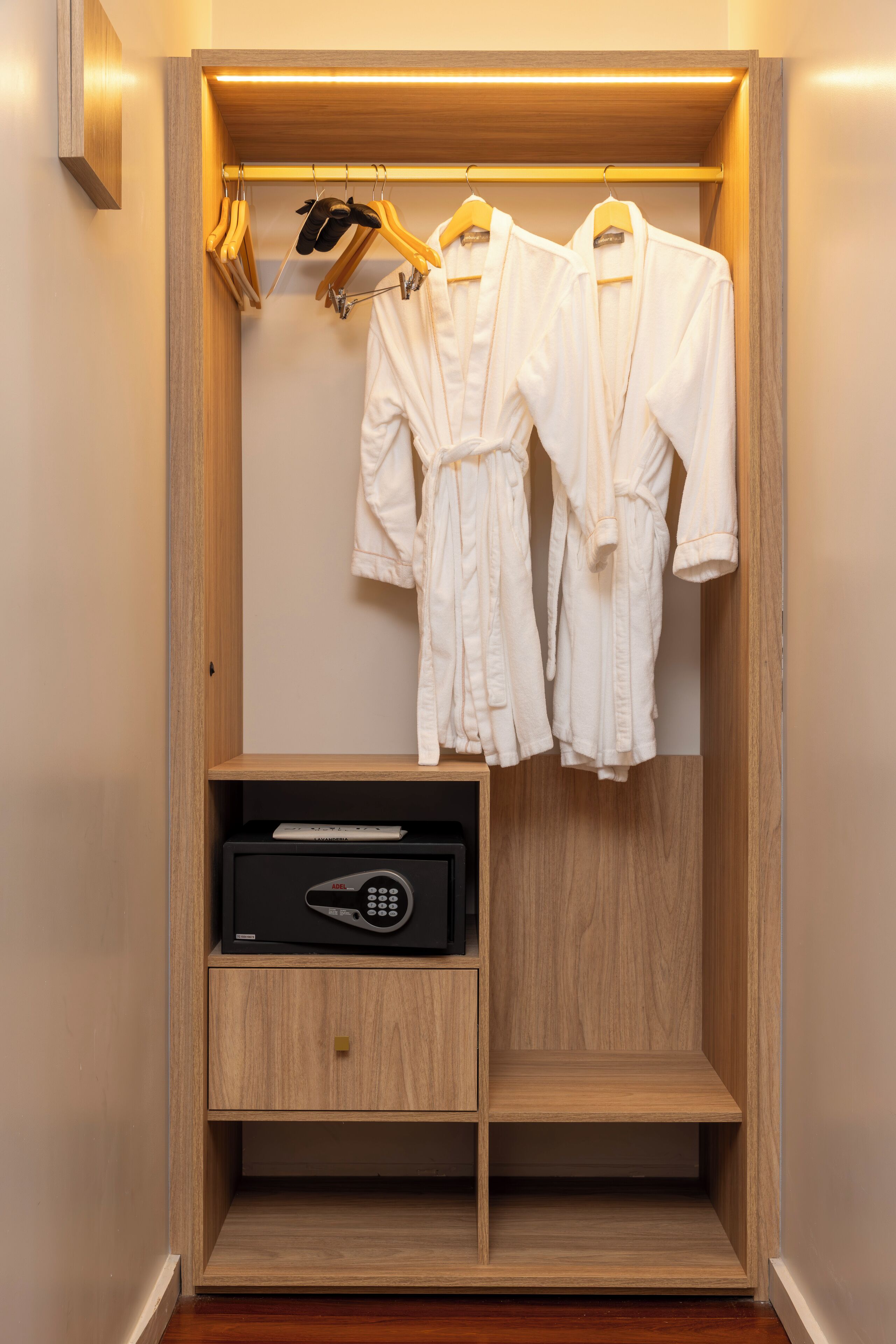 A wooden walk-in closet with two white robes hanging on a rack, a black microwave, and a small drawer unit.