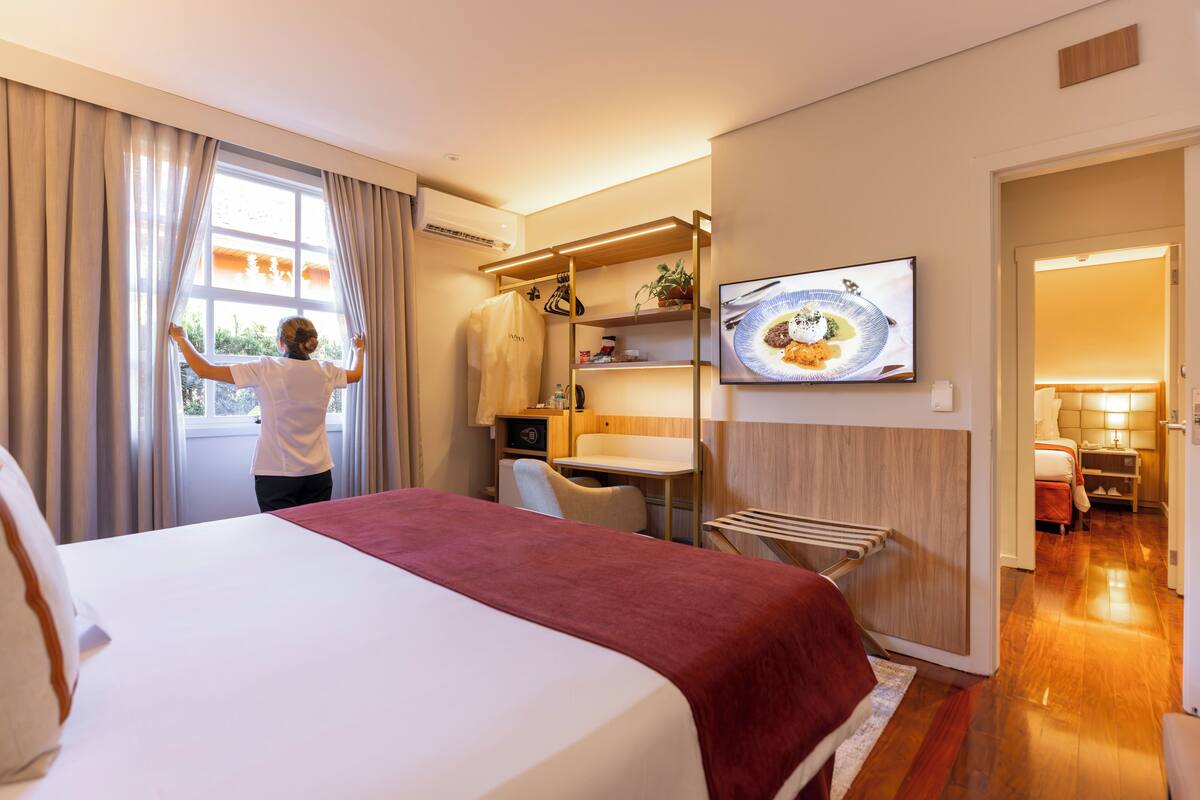 Quarto de hotel com cama, escrivaninha, cadeira, televisão e janela com cortinas.