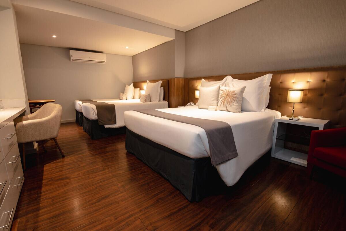 Quarto de hotel com três camas, uma escrivaninha, uma cadeira e uma mesa de cabeceira com abajur.