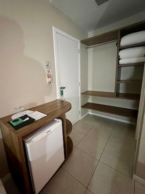 A compact hotel room with a wooden desk, a mini fridge, a white door, and a built-in wardrobe with shelves.