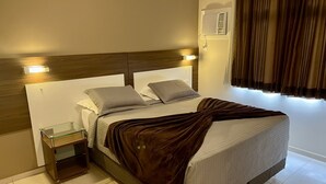 A neatly made bed with a brown blanket, a bedside table with a glass top, and a wall-mounted air conditioning unit. - Brisamar Suite Hotel (Florianópolis)