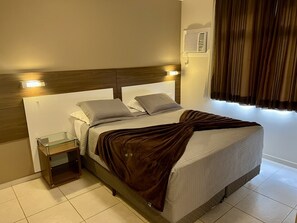 Uma cama bem arrumada com um cobertor marrom, uma mesa de cabeceira com tampo de vidro e um ar condicionado fixado na parede.