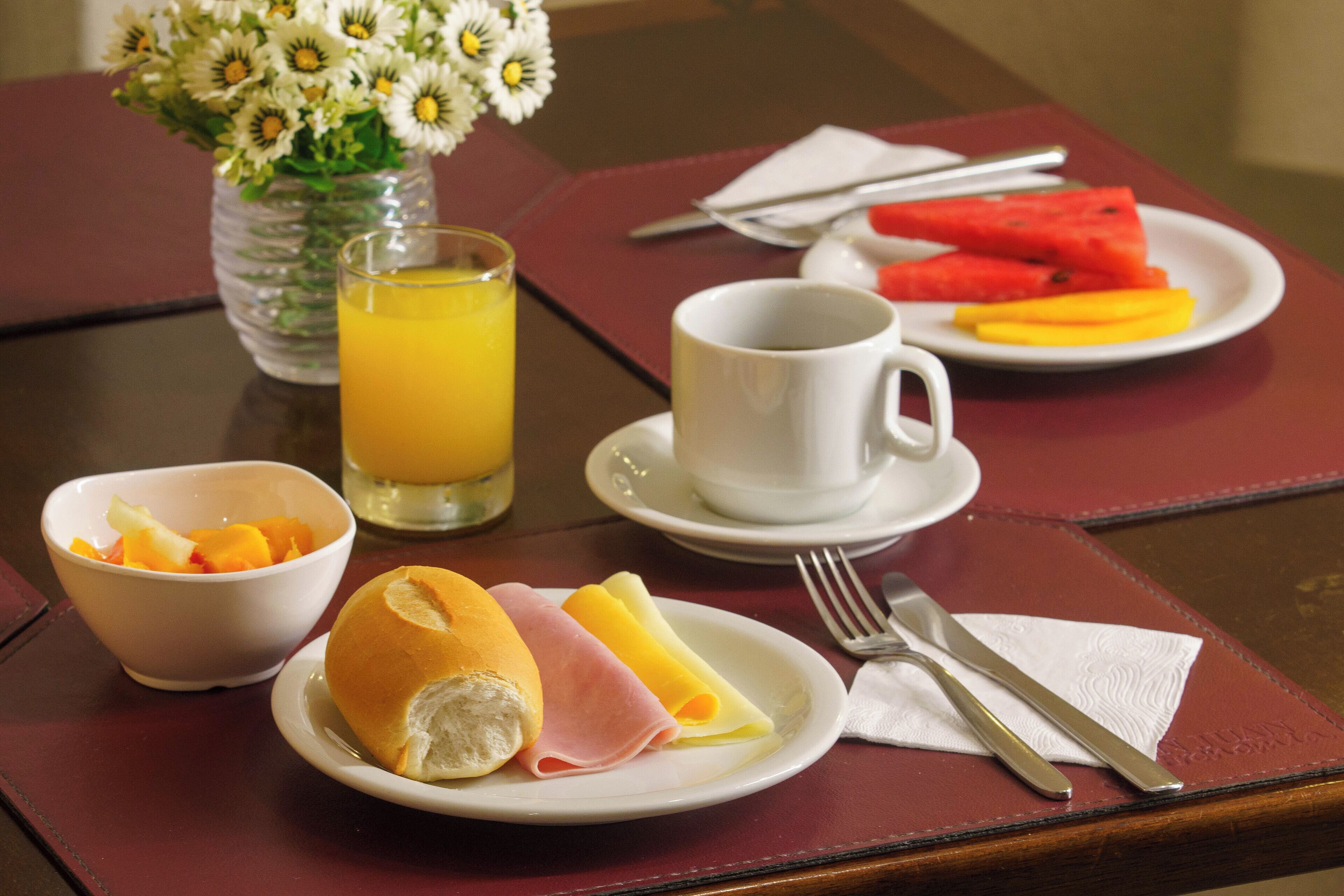 Mesa posta para o café da manhã, com um prato de ovos, presunto, frutas, um copo de suco de laranja e um vaso de flores.