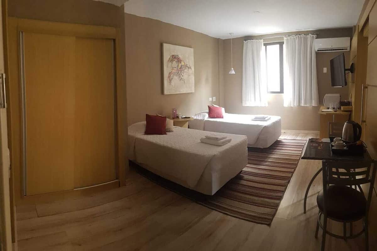 Een hotelkamer met twee bedden, een bureau, een stoel, een raam met gordijnen en een schilderij aan de muur.
