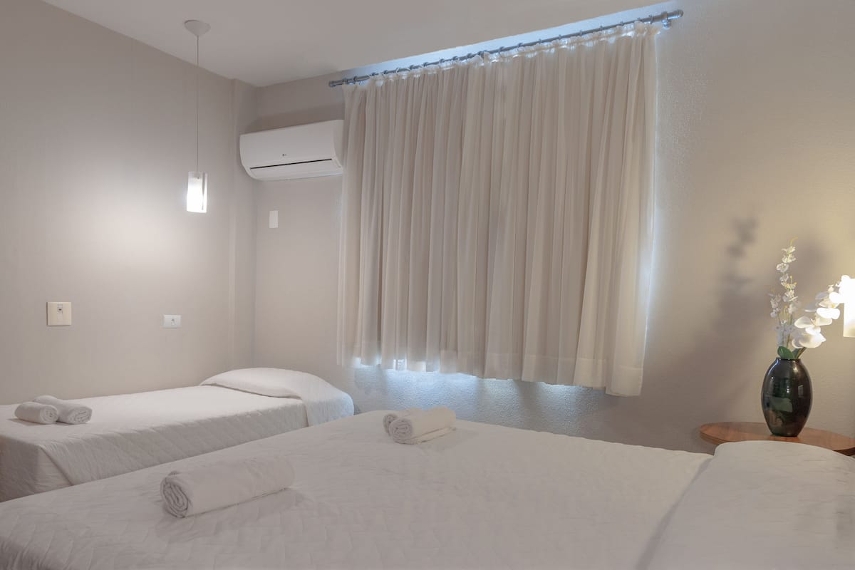 Een hotelkamer met twee bedden, een raam met gordijnen, een airconditioning en een bloemenvaas op een nachtkastje.