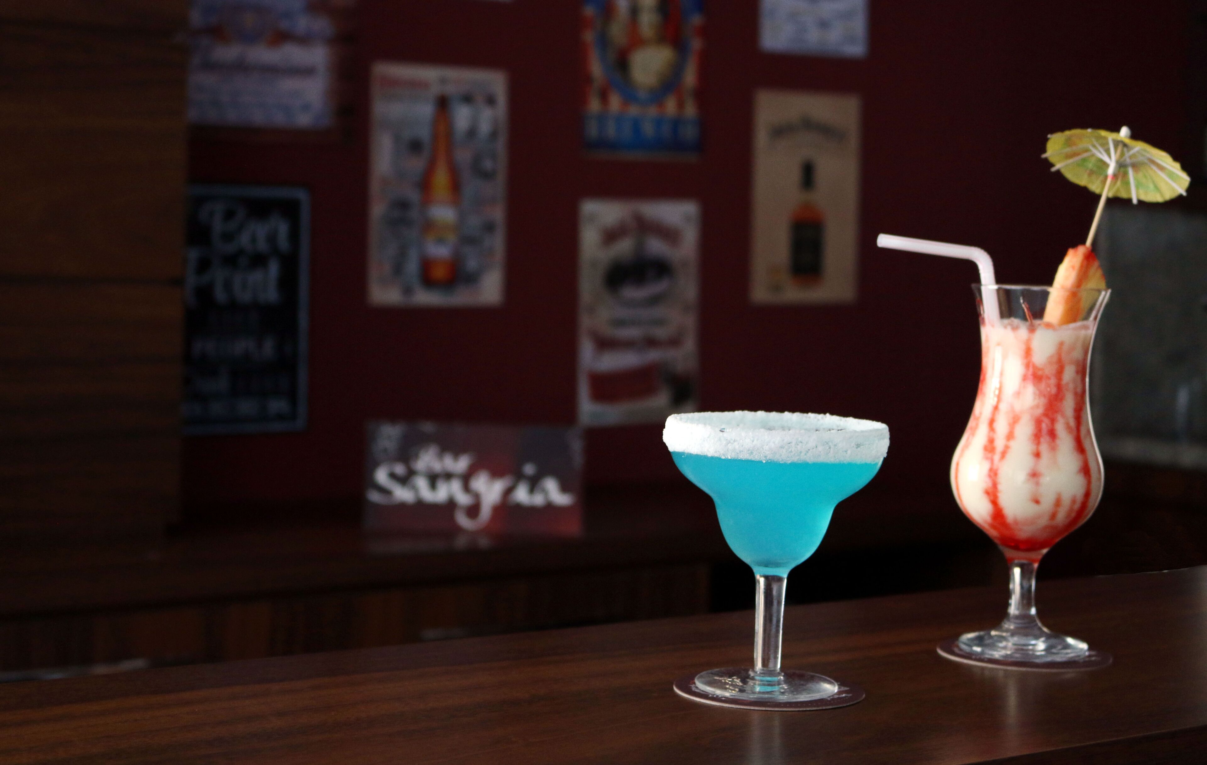 Two cocktails on a bar counter with a blurred background of posters and signs.