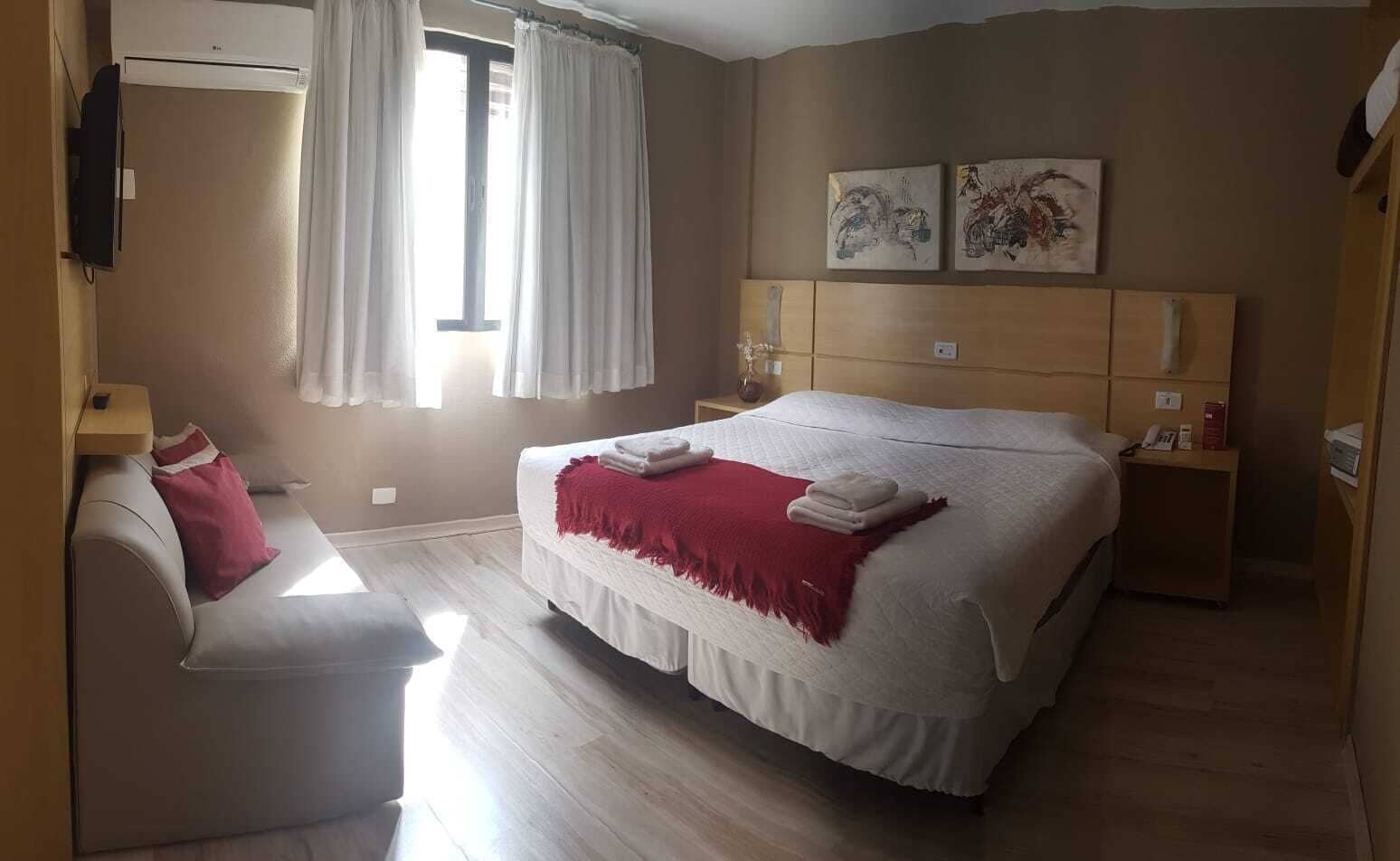 A bedroom with a bed, a bedside table, a nightstand, a chair, and two wall-mounted paintings.