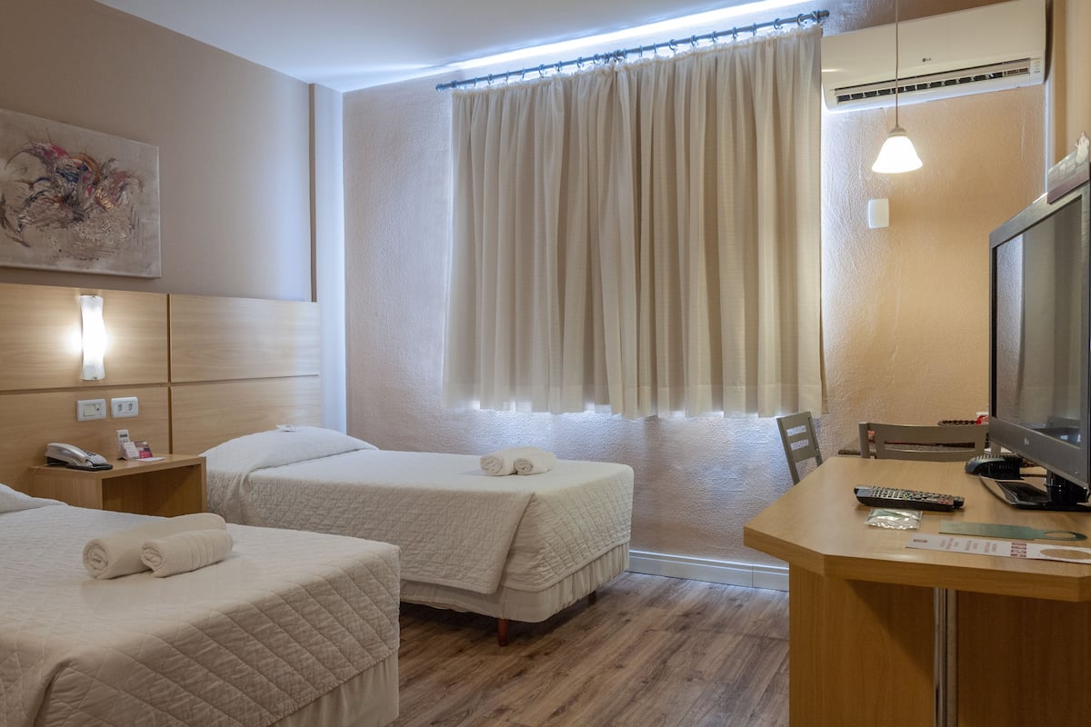 Een hotelkamer met twee bedden, een bureau met een computer, een stoel en een raam met gordijnen.