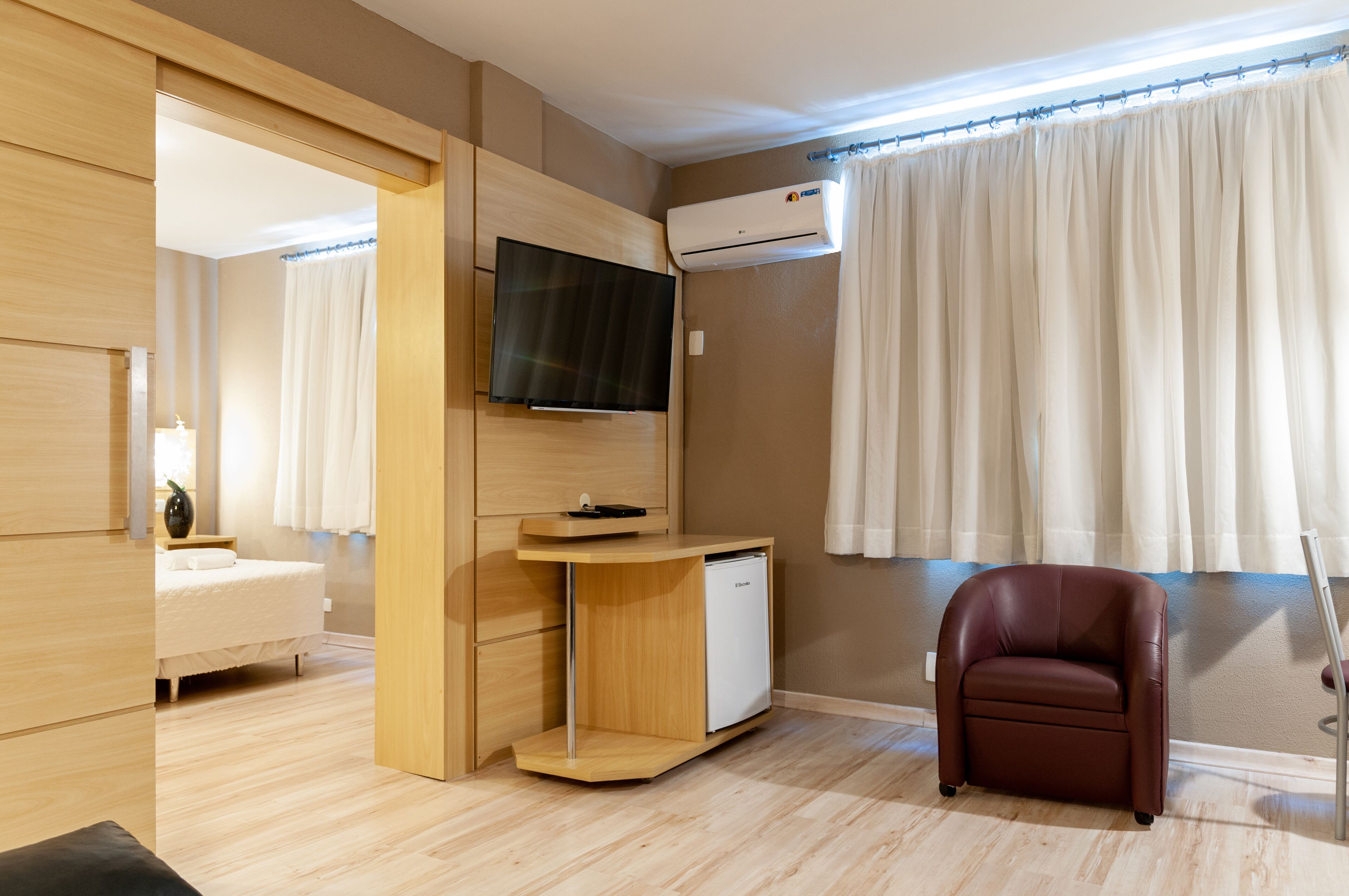Una habitación de hotel moderna con una cama, un televisor montado en la pared, un refrigerador blanco pequeño y un sillón de cuero.